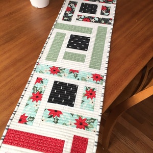 Puede incluir: Un camino de mesa blanco con un diseño de patchwork con cuadrados y rectángulos rojos, verdes y negros. El camino de mesa tiene un fondo blanco con un borde rayado gris y blanco.