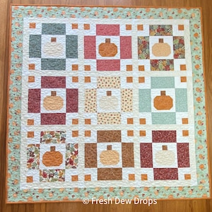 May include: A quilt with a patchwork design featuring various fall-themed fabrics. The quilt is made up of nine squares, each with a different pattern. The squares are arranged in a 3x3 grid. The quilt is bordered with a light green fabric with a floral pattern. The quilt is on a wooden floor. © Fresh Dew Drops