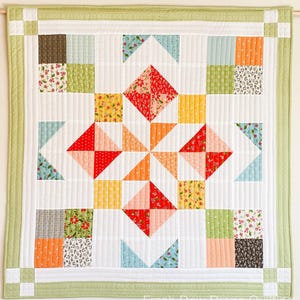 Può includere: Una trapunta bianca con un bordo verde e un design a patchwork colorato. La trapunta presenta un motivo centrale a stella con quadrati e triangoli rossi, arancioni, gialli e blu. Il bordo esterno della trapunta è fatto di quadrati più piccoli in vari colori e motivi.