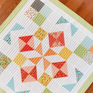 May include: A white quilt with a starburst pattern in the center. The quilt is made with various floral fabrics in shades of red, orange, yellow, green, and blue. The quilt is edged with a light green border.