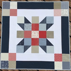 May include: A red, white, and blue patchwork quilt with a starburst design in the center. The quilt is made of various fabrics with different patterns, including gingham, polka dots, and stripes. The quilt is framed with a navy blue border.