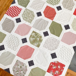 May include: A quilt with a geometric pattern featuring hexagons in various shades of green, red, and white. The quilt is made with a variety of fabrics with different patterns, including floral, polka dot, and striped. The quilt is finished with a black border.