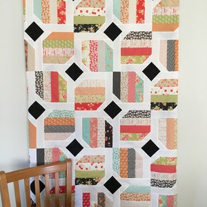 May include: A white quilt with black diamond shapes and colourful fabric squares. The quilt is hanging on a white wall.