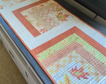 Peut inclure: Un chemin de table matelassé sur le thème de l'automne. Le chemin de table présente un design en patchwork avec différentes nuances d'orange, de brun, de vert et de blanc. Le tissu est imprimé de feuilles et des mots "Automne" et "Chêne écarlate".