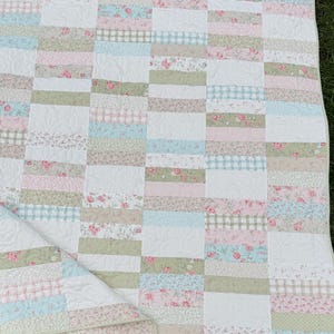 Op de afbeelding: Een patchwork quilt met een herhalend patroon van rechthoekige stoffen stroken in zachte pasteltinten zoals roze, saliegroen en lichtblauw. Het ontwerp bevat bloemenprints, ruitjespatronen en witte secties met doorgestikte krulmotieven.