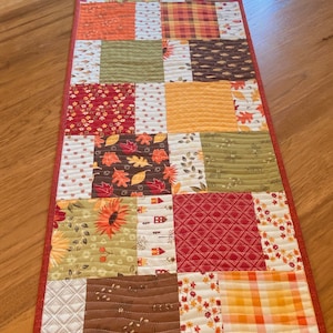 May include: A patchwork table runner with a variety of fall-themed fabrics in shades of red, orange, yellow, green, and brown. The runner is quilted and has a red border.