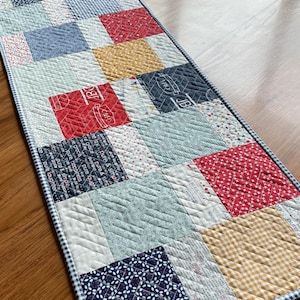 Puede incluir: Un camino de mesa de patchwork con una variedad de patrones y colores, incluyendo rojo, azul, amarillo y blanco. El camino de mesa está acolchado con un patrón de diamante y tiene un borde de vichy blanco y azul.