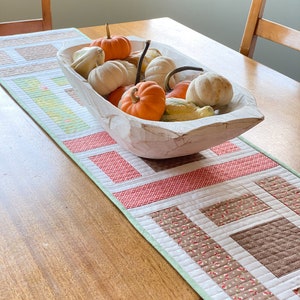 May include: A white wooden bowl filled with gourds and small pumpkins, sitting on a table runner with a patchwork design of brown, red, and white fabric. The runner has a green border.