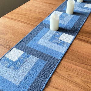 Könnte beinhalten: Ein blauer und weißer gesteppter Tischläufer mit geometrischem Muster. Vier weiße Kerzen stehen auf dem Läufer, der auf einem Holztisch liegt.