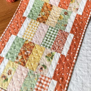 Peut inclure: Un chemin de table matelassé rectangulaire présentant un motif patchwork de bandes de tissu verticales dans des tons orange, jaune, vert et blanc. Les tissus incluent des motifs floraux, des carreaux vichy et des pois, avec une bordure matelassée blanche.