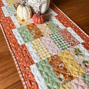 May include: A quilted table runner featuring a patchwork design of rectangular fabric blocks in shades of orange, yellow, green, and white with floral and gingham patterns. Three decorative pumpkins sit on top. The text Fresh Dew Drops is visible.