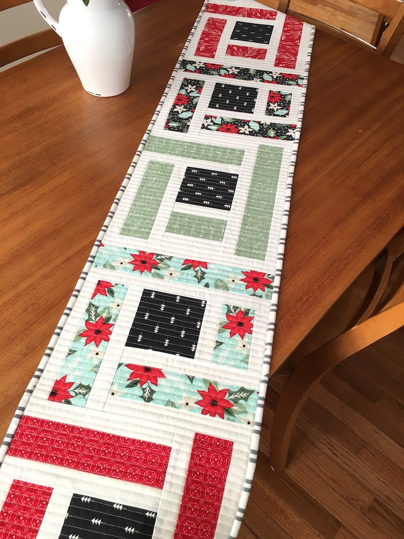 K&ouml;nnte beinhalten: Ein wei&szlig;er Tischl&auml;ufer mit Patchwork-Design mit roten, gr&uuml;nen und schwarzen Quadraten und Rechtecken. Der L&auml;ufer hat einen wei&szlig;en Hintergrund mit einem grau-wei&szlig;en gestreiften Rand.