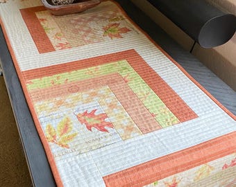 May include: A quilted table runner with a fall leaf design in shades of orange, green, and brown. The runner features a white background with a quilted pattern and a border of orange fabric. The text "SCARLET OAK" is visible on the runner.