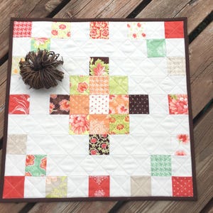 May include: A square, quilted patchwork table topper with a white background and a brown border. The quilt features a cross design made of colourful floral and patterned squares in shades of red, orange, green, and brown. A decorative brown ball sits on the quilt.