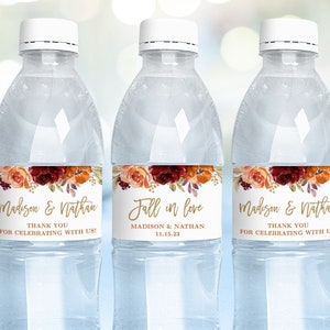 May include: Three clear plastic water bottles with labels featuring a floral design in shades of red, orange, and gold. The labels also include the text "Madison & Nathan" and "Fall in love" with the date "11.15.23".