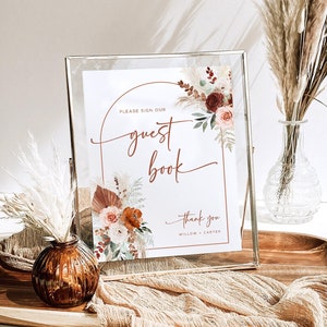 May include: A framed guest book sign with floral accents in shades of rust, peach, and burgundy. The sign reads "Please Sign Our Guest Book" and "Thank You" with names below. The frame is clear with a gold-colored metal stand.