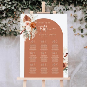 Peut inclure: Un plan de table de couleur terre cuite avec un motif floral et le texte "Find Your Table" en écriture blanche. Le plan de table liste les noms des invités pour chaque numéro de table, avec les noms de la mariée et du marié en haut : "Willow + Carter".