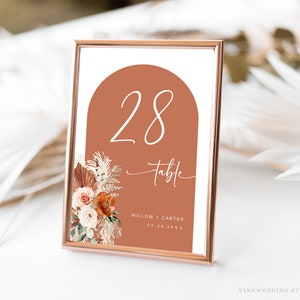 May include: A terracotta-colored arch with the number 28 and the word "table" in white script. The arch is surrounded by a floral arrangement with dried grasses and flowers in shades of white, orange, and brown. The text "Willow + Carter 09.22.20XX" is printed below the arch.