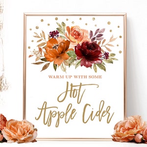 Puede incluir: Un letrero enmarcado con un arreglo floral de flores naranjas, rojas y burdeos. El letrero dice "Warm up with some Hot Apple Cider" en escritura dorada. Puntos de purpurina dorada est&aacute;n dispersos en la parte superior del fondo blanco.