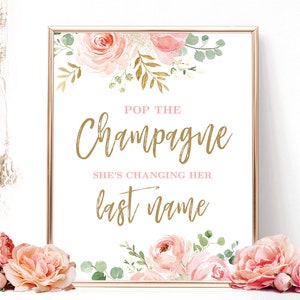 May include: A printable sign with pink and gold floral illustrations. The text reads "Pop the Champagne She's Changing Her Last Name".