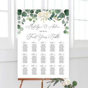 May include: A white wedding seating chart with green eucalyptus and white roses at the top. The chart is titled "Find Your Table" and lists the names of guests assigned to each table number.