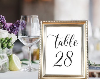 Op de afbeelding: Een gouden gelijst tafelnummerbord met een witte achtergrond en zwarte tekst. Het bord heeft de tekst "Tafel 28" in een cursief schrift.