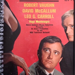May include: A vintage poster for the TV show "The Man From U.N.C.L.E." starring Robert Vaughn and David McCallum. The poster features a red background with the show's title in white letters. The actors' names are listed below the title in smaller white letters. The poster also includes the show's credits, including the name of the arranger and conductor, Hugo Montenegro.