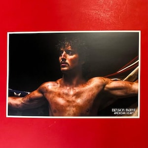 May include: A photograph of a man with a muscular physique, holding a portion of an American flag. The image has a dark background and the text "BENSON BOONE AMERICAN HEART" is visible in the lower right corner.