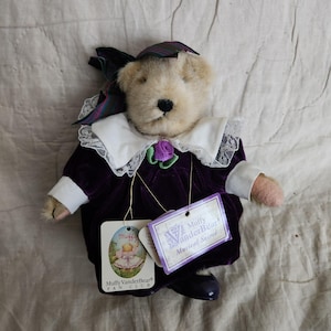 May include: A plush teddy bear wearing a purple velvet dress with a white lace collar and a matching bow. The bear has a purple rose accent and is holding two tags that read "Muffy VanderBear". The bear is wearing black shoes.