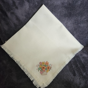 May include: Cream-colored, square cross-stitch napkin with a fringed edge. The napkin features a floral design in a small pot, with various colors including pink, yellow, and orange. The napkin is folded diagonally.