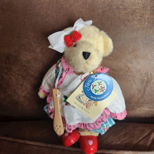 May include: A plush teddy bear wearing a white bow with red strawberry accents, a pink and white apron, and red shoes with strawberry details. The bear holds a wooden spoon and has a tag that reads "Jam Session Muffy VanderBear".