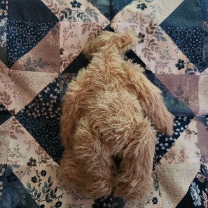 May include: A brown plush teddy bear with long, shaggy fur is lying on a patchwork quilt with a floral pattern. The quilt is made of different fabrics in shades of brown, beige, and black.