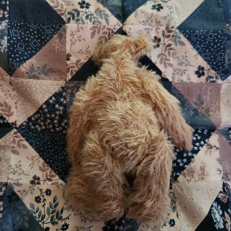 May include: A brown plush teddy bear with long, shaggy fur is lying on a patchwork quilt with a floral pattern. The quilt is made of different fabrics in shades of brown, beige, and black.