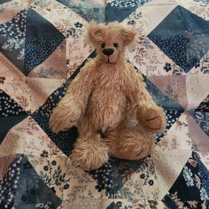May include: A brown teddy bear with a fluffy coat sits on a patchwork quilt with a variety of floral patterns in shades of blue, brown, and cream.