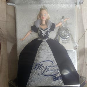 May include: A blonde doll wearing a dark blue and silver gown with a high collar, a silver tiara, and a necklace. The doll is inside a clear display box with a silver ornament labeled Millennium Keepsake and the text Millennium Princess Barbie.
