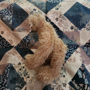 May include: A brown teddy bear with long, shaggy fur is sitting on a patchwork quilt with a floral pattern. The quilt is made of various shades of blue, brown, and white fabric.