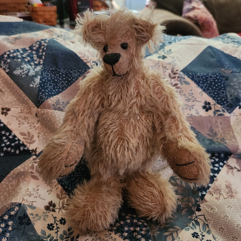 May include: A light brown teddy bear with a fluffy texture, sitting on a patterned quilt. The bear has a long snout and black eyes.