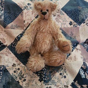 May include: A light brown teddy bear with a fluffy texture sits on a patchwork quilt with a floral pattern. The quilt is made of various shades of blue, brown, and white.