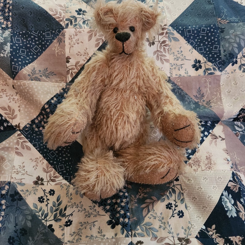 May include: A light brown teddy bear with a fluffy texture sits on a patchwork quilt with a floral pattern. The quilt is made of various shades of blue, brown, and white.