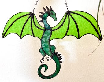 May include: A stained glass dragon with outstretched wings. The dragon is primarily green with black outlines and a chain for hanging. The wings are a bright, translucent green, and the body is a darker shade of green with lighter green spots.