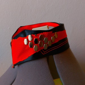 Peut inclure: Un collier ras de cou noir et rouge avec des clous hexagonaux argentés. Le collier est fait d'un tissu en maille et a une bordure en cuir noir.