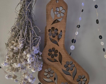 May include: Two wooden sock-shaped ornaments with floral cutouts hang from a wooden beam. The ornaments are a warm brown color and have a decorative design. Dried flowers and a string of buttons are also visible.