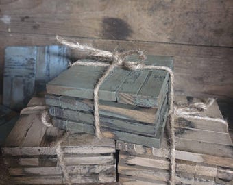May include: Stacks of square wooden coasters, some painted in muted green and others in natural wood tones. Each stack is bound with twine, creating a rustic and handcrafted aesthetic. The coasters appear to be made from reclaimed wood.