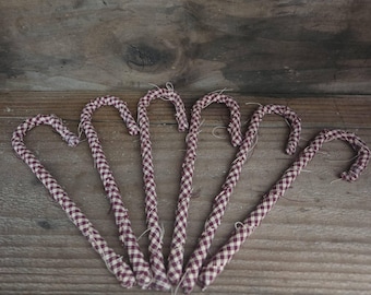May include: A set of five candy cane ornaments wrapped in a maroon and white gingham fabric. Each ornament features a curved top and a straight stem, with visible frayed fabric edges and loose threads along the length of the candy cane shapes.