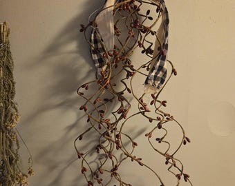 May include: A decorative hanging bundle of thin, twisted brown branches adorned with small, dark red berries. The arrangement is tied at the top with a dark blue and white checkered fabric ribbon and features a single rusted metal bell accent.