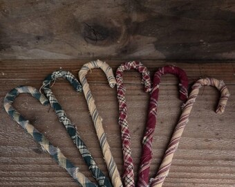 May include: Six candy cane ornaments wrapped in various fabric strips. The patterns include blue and tan plaid, solid tan, red and white plaid, solid dark red, and tan with red stripes. Each ornament features frayed fabric edges and a curved top.