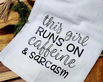 May include: White kitchen towel with black text that reads "This girl runs on caffeine & sarcasm".