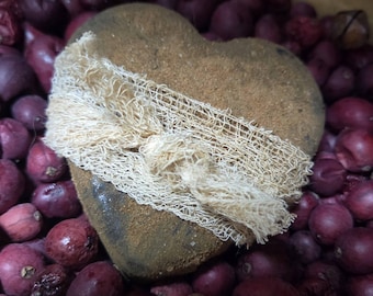 May include: A heart-shaped stone with a rough, textured surface, wrapped with a beige, open-weave fabric. The stone is surrounded by small, dark red, round objects, creating a rustic, natural aesthetic.