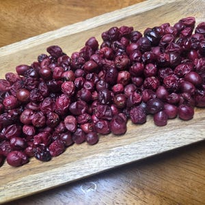 May include: A wooden tray filled with a large pile of dried, dark red berries. The berries are various sizes and shapes, with a wrinkled texture. The tray is made of light-colored wood, and the berries are the focal point of the image.