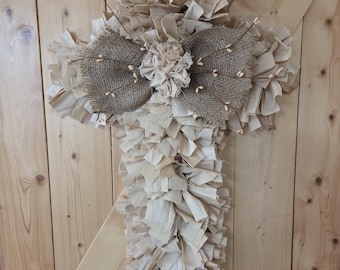 May include: A cross-shaped wall hanging constructed from numerous strips of light beige fabric tied to a frame. The center features a textured brown burlap bow adorned with a fabric flower and thin twigs with small, light-colored decorative buds.